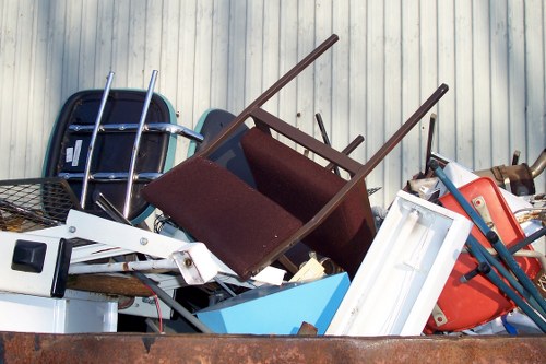 A large pile of assorted discarded household items and furniture stacked in an outdoor area, possibly on a driveway or nearby land. The collection includes a damaged wooden chair with a dark red fabric seat, a blue plastic storage container, a white plastic food storage box, an orange plastic item, and a black wire basket. Some metal legs and parts of furniture are visible among the debris. The background features vertical metallic or corrugated paneling, creating a neutral backdrop that highlights the bulk of rubbish in the foreground. The items appear to be a mixture of broken or unwanted household objects awaiting collection or removal, consistent with rubbish clearance activities. The scene suggests an environment prepared for a professional house clearance or waste disposal service, such as those provided by Haringey House Clearance, operating in the local Haringey area, possibly near London. The debris is piled in a somewhat disorganized manner, emphasizing the nature of rubbish collection and clearance work commonly undertaken in urban and suburban settings involving rubbish removal from domestic properties.